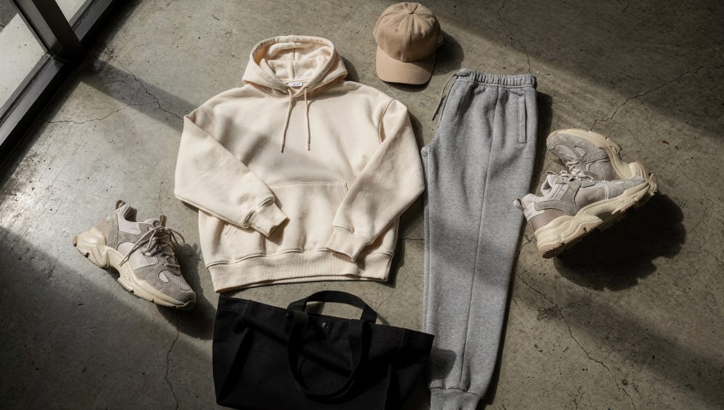 Tenue streetwear posée au sol : hoodie oversize crème, jogging gris, casquette beige, sneakers et tote bag noir