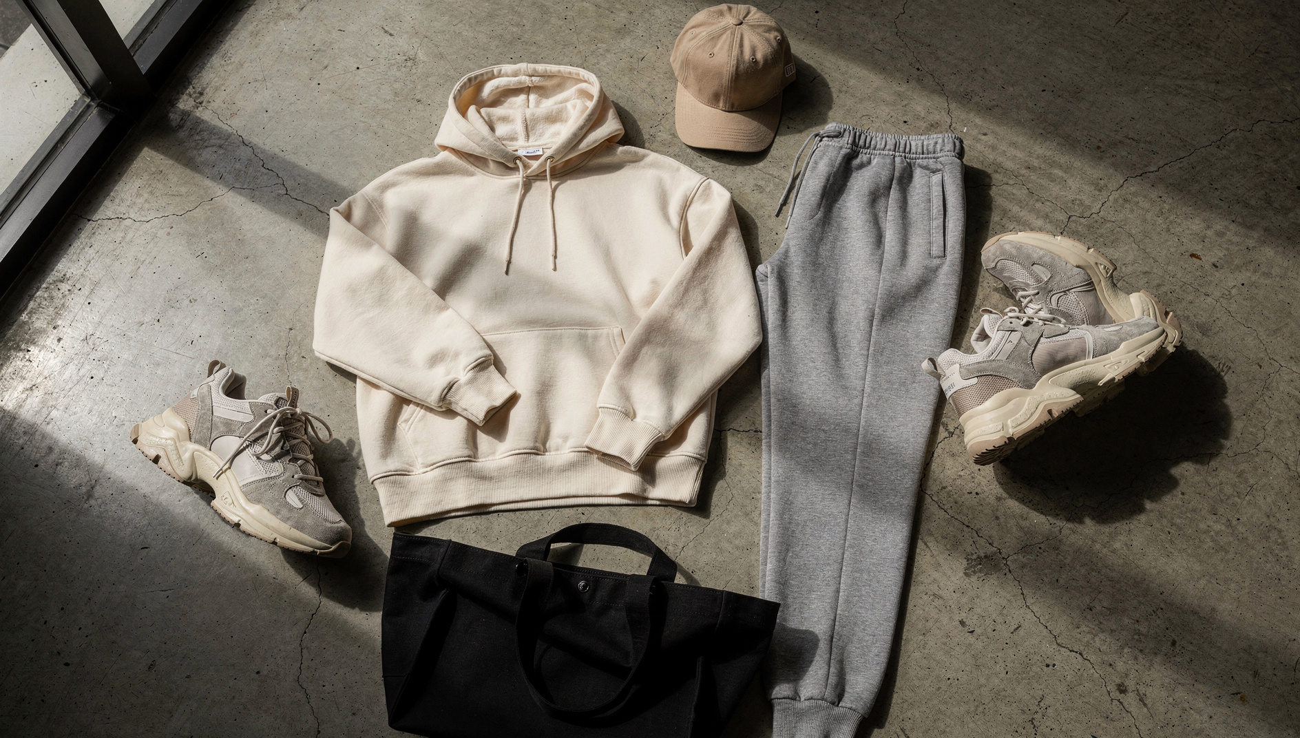Tenue streetwear posée au sol : hoodie oversize crème, jogging gris, casquette beige, sneakers et tote bag noir