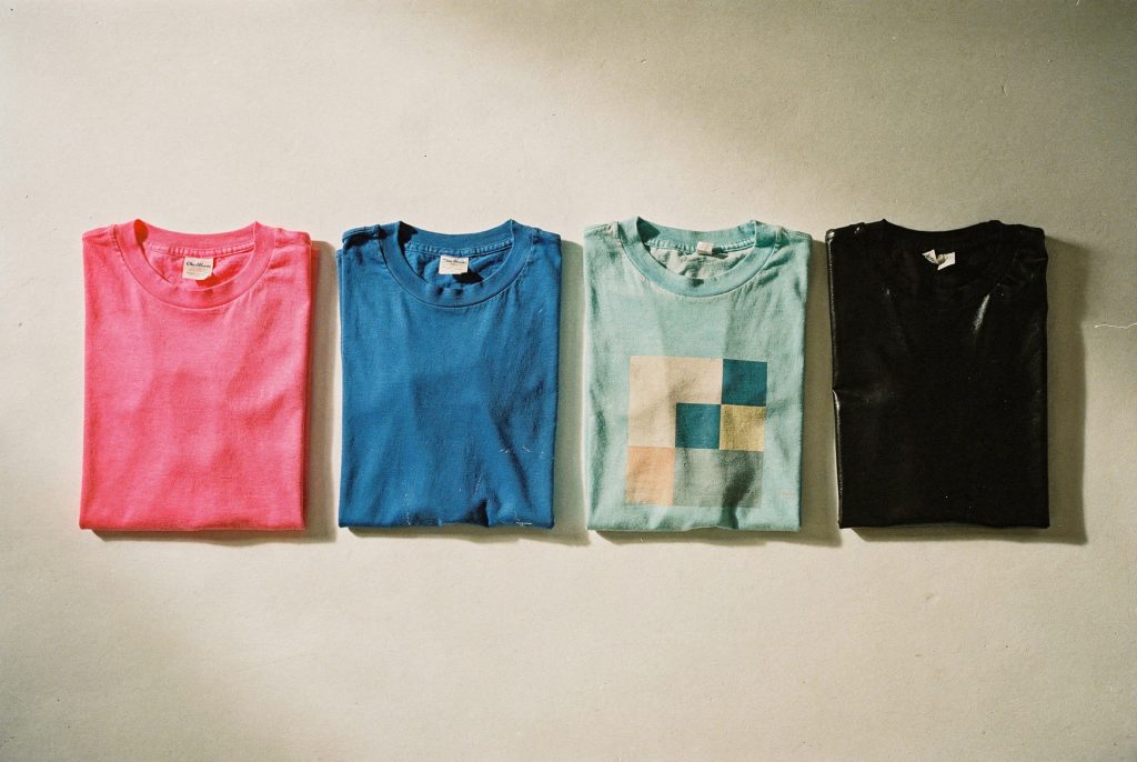 Quatre tee-shirts des années 80 pliés et alignés sur fond écru : rose fluo, bleu électrique, turquoise à motif géométrique et noir